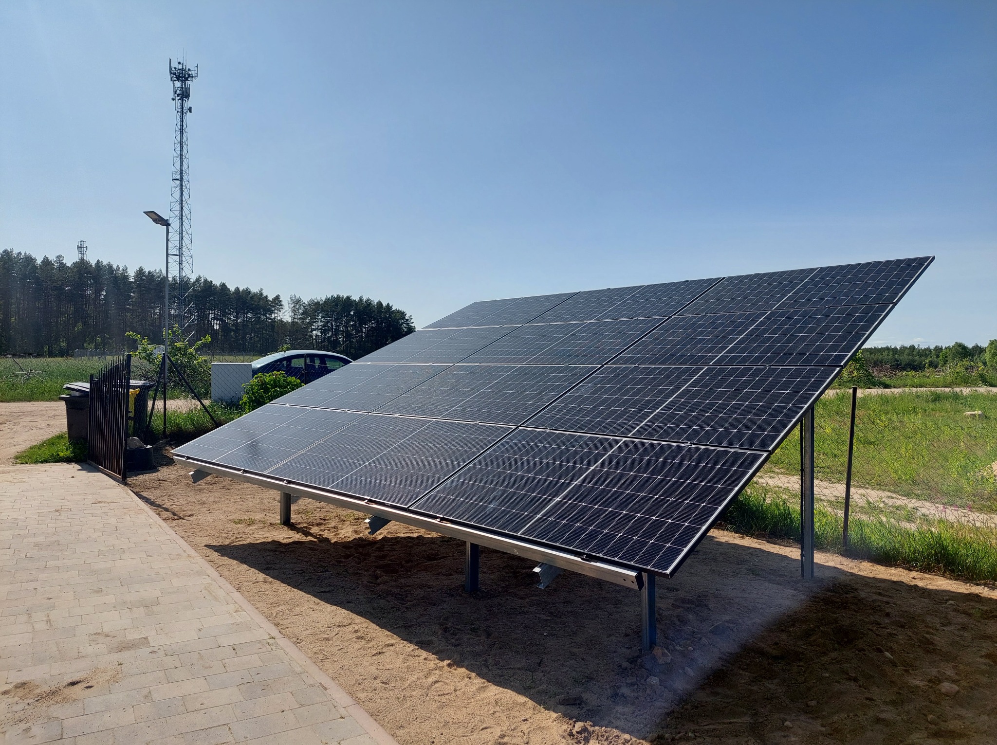 Panel fotowoltaiczny na metalowej konstrukcji, ułożony na piaszczystym podłożu, obok chodnika z kostki. W tle las i wieża telekomunikacyjna.