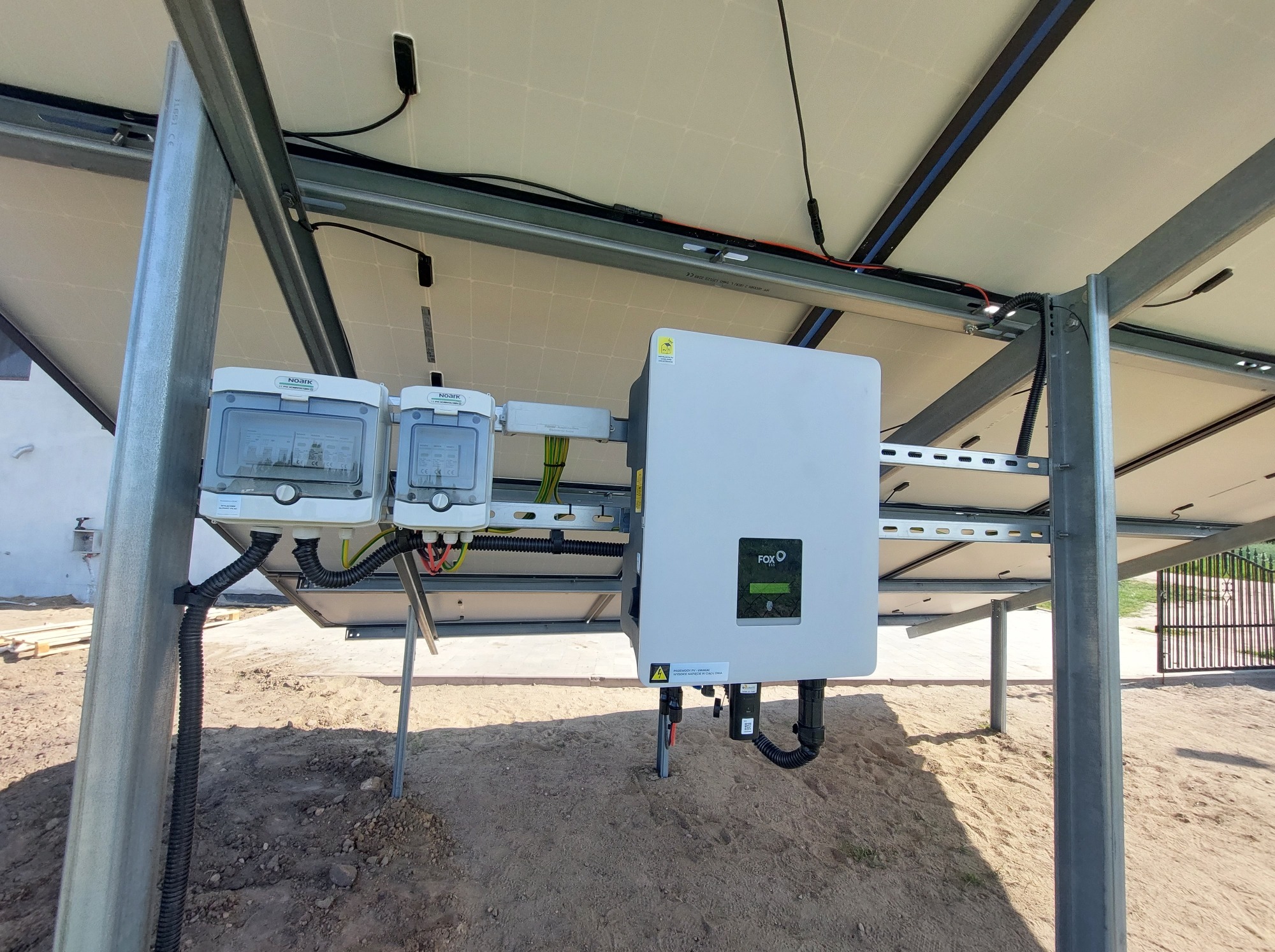Fotowoltaika: Inwerter i zabezpieczenia na konstrukcji wsporczej, widok z dołu. Instalacja paneli słonecznych na gruncie, szara ziemia w tle.
