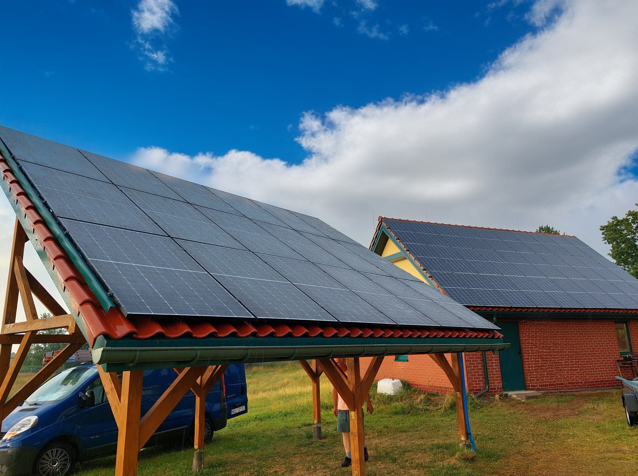 Instalacja paneli słonecznych na wiacie i budynku z czerwonej cegły, błękitne niebo z chmurami w tle. Widoczny samochód dostawczy.