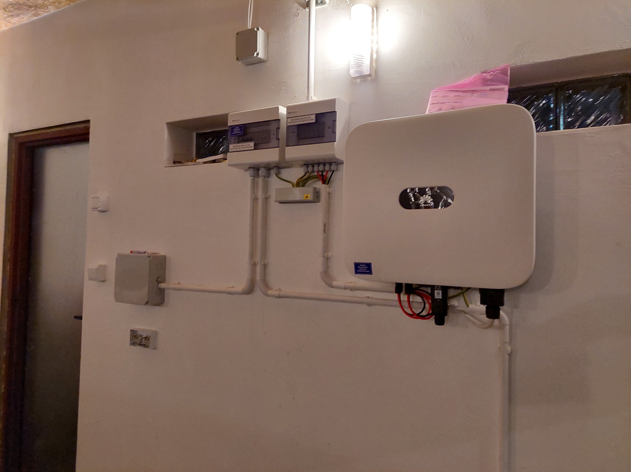 Zamontowany inwerter fotowoltaiczny na ścianie wewnątrz budynku, z widocznymi przewodami i zabezpieczeniami. Instalacja elektryczna w pomieszczeniu.