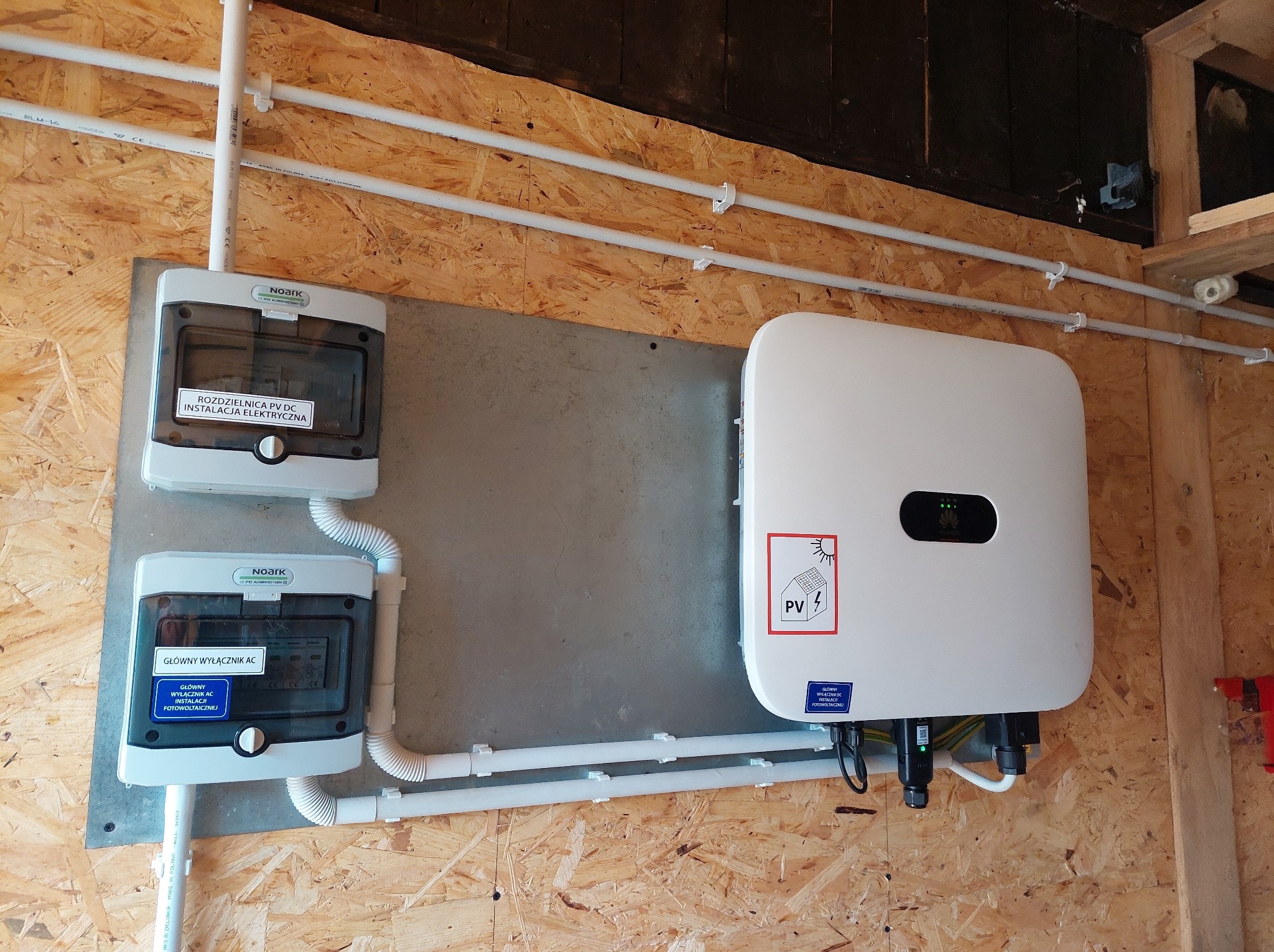 Kompleksowa instalacja fotowoltaiczna wewnątrz budynku: falownik, rozdzielnica PV DC i wyłącznik AC, połączone białymi rurami. Montaż na płycie OSB.