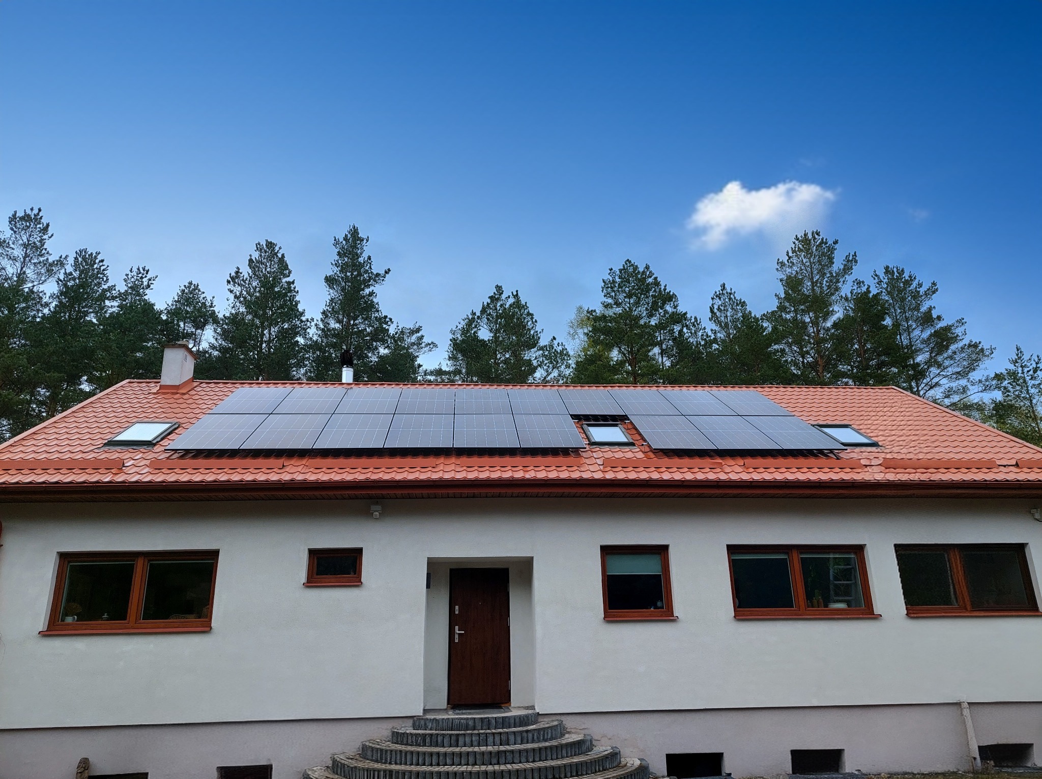 Dom jednorodzinny z panelami fotowoltaicznymi na czerwonym dachu, widok z dołu, błękitne niebo z pojedynczą chmurą, drzewa w tle.