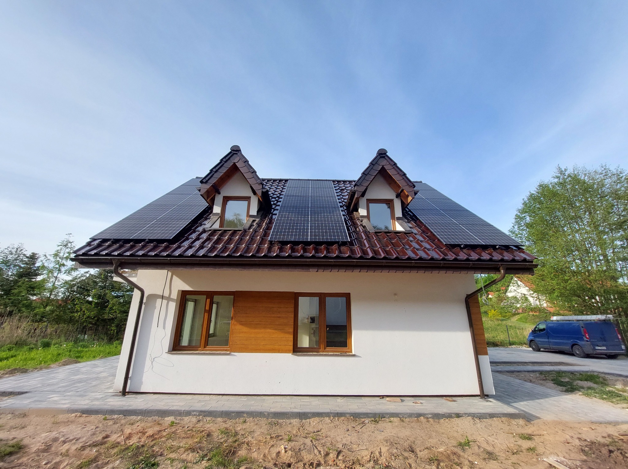 Dom jednorodzinny z panelami fotowoltaicznymi na dachu pokrytym czerwoną dachówką. Błękitne niebo w tle. Widoczny fragment chodnika i zaparkowany samochód.
