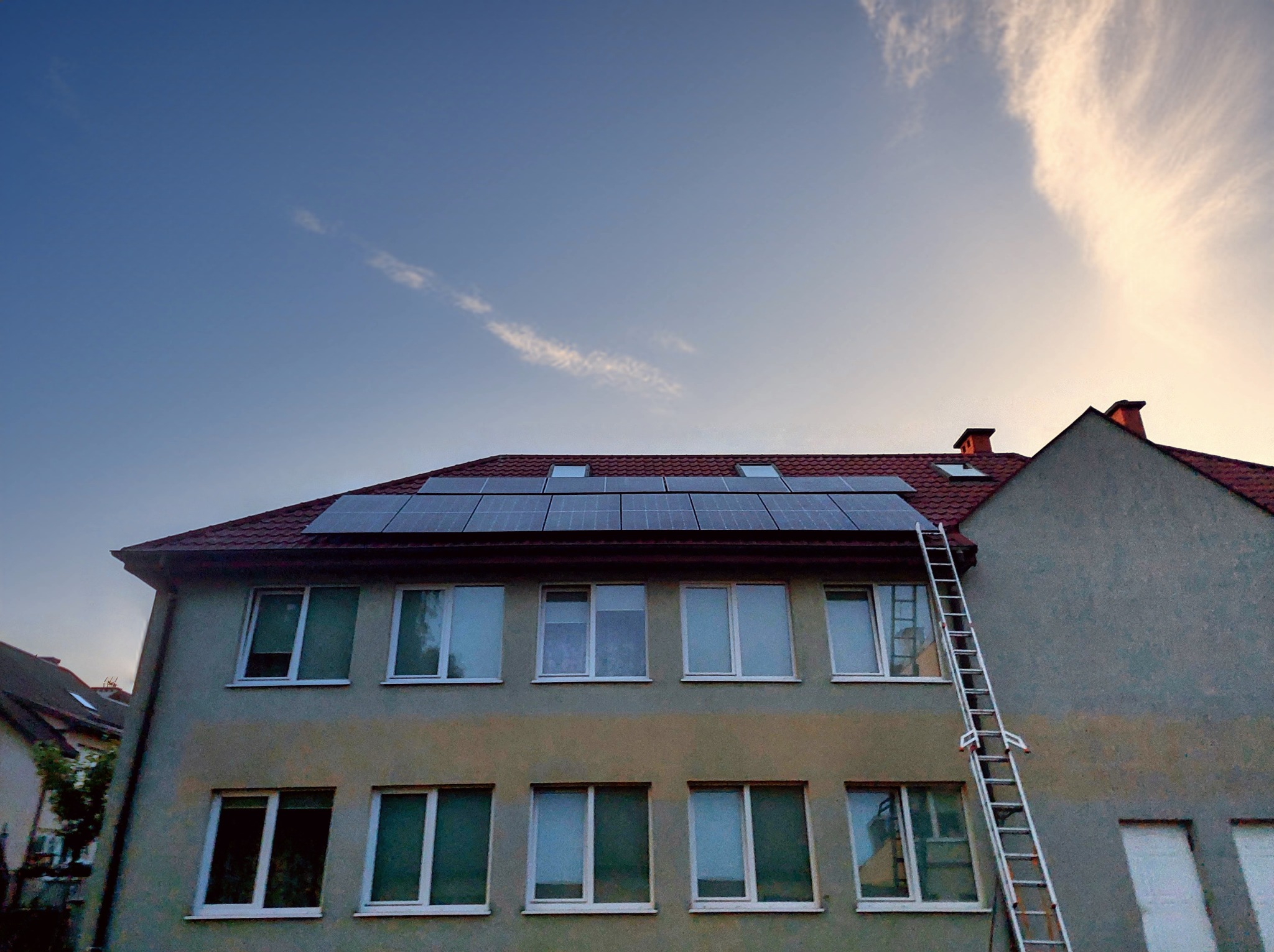 Panele słoneczne na dachu budynku z czerwoną dachówką, drabina oparta o ścianę, błękitne niebo z chmurami w tle. Widok z dołu.