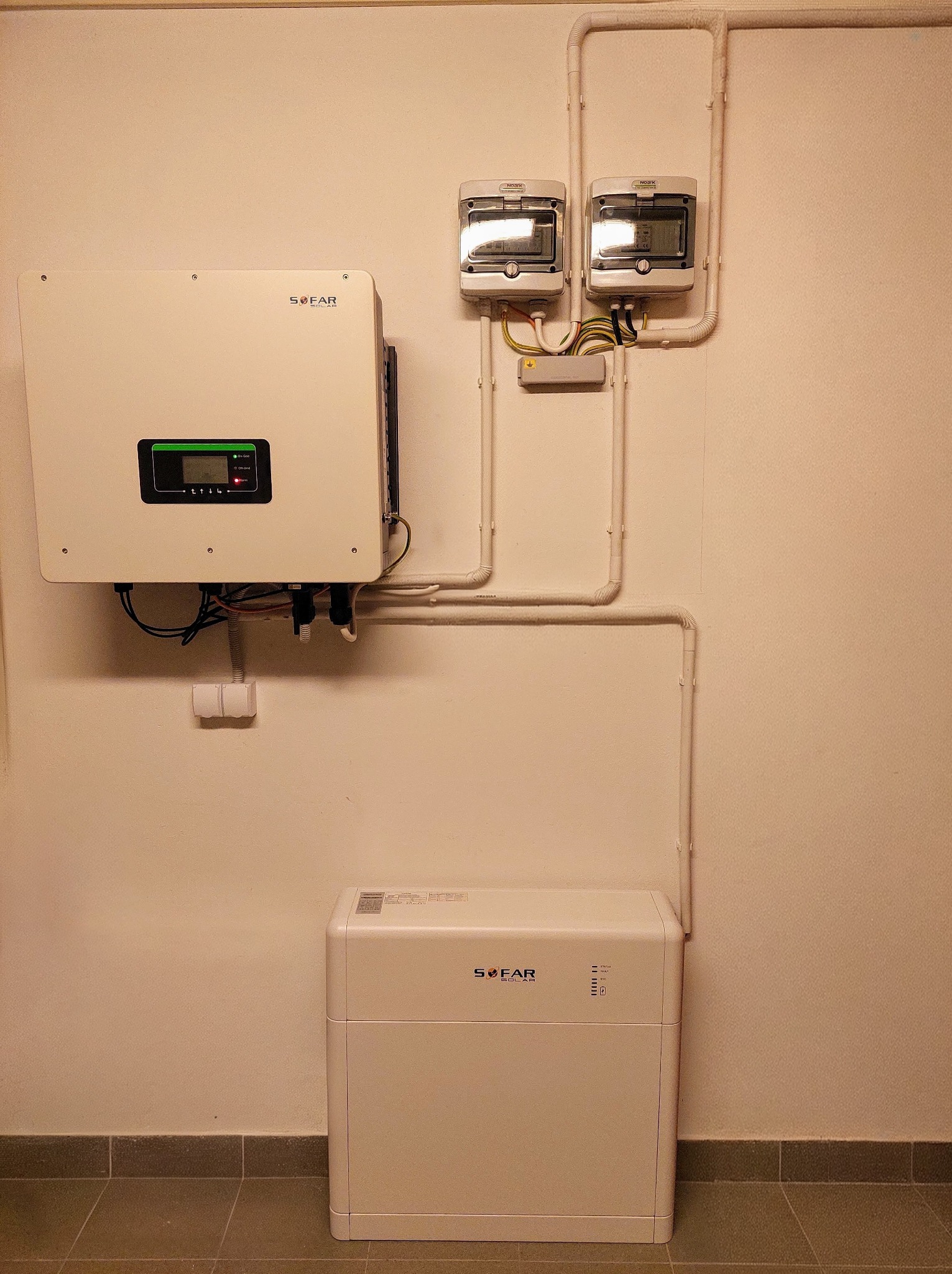 Biały inwerter SOFAR, licznik energii i akumulator na ścianie w pomieszczeniu. Widoczne przewody i instalacja elektryczna. Całość tworzy system fotowoltaiczny.