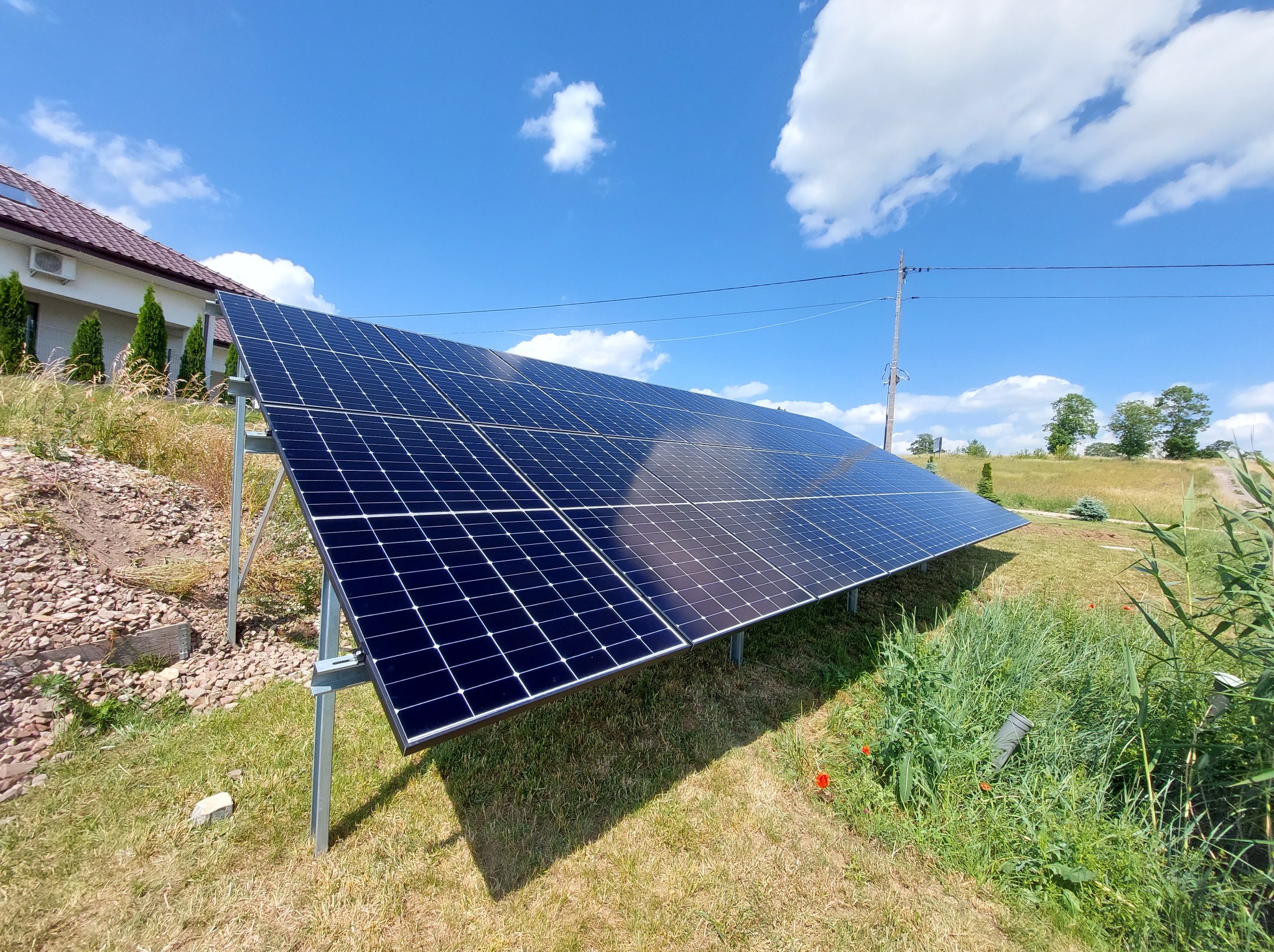 Panel fotowoltaiczny na metalowej konstrukcji, zainstalowany na trawie obok domu z czerwoną dachówką, na tle błękitnego nieba z chmurami.