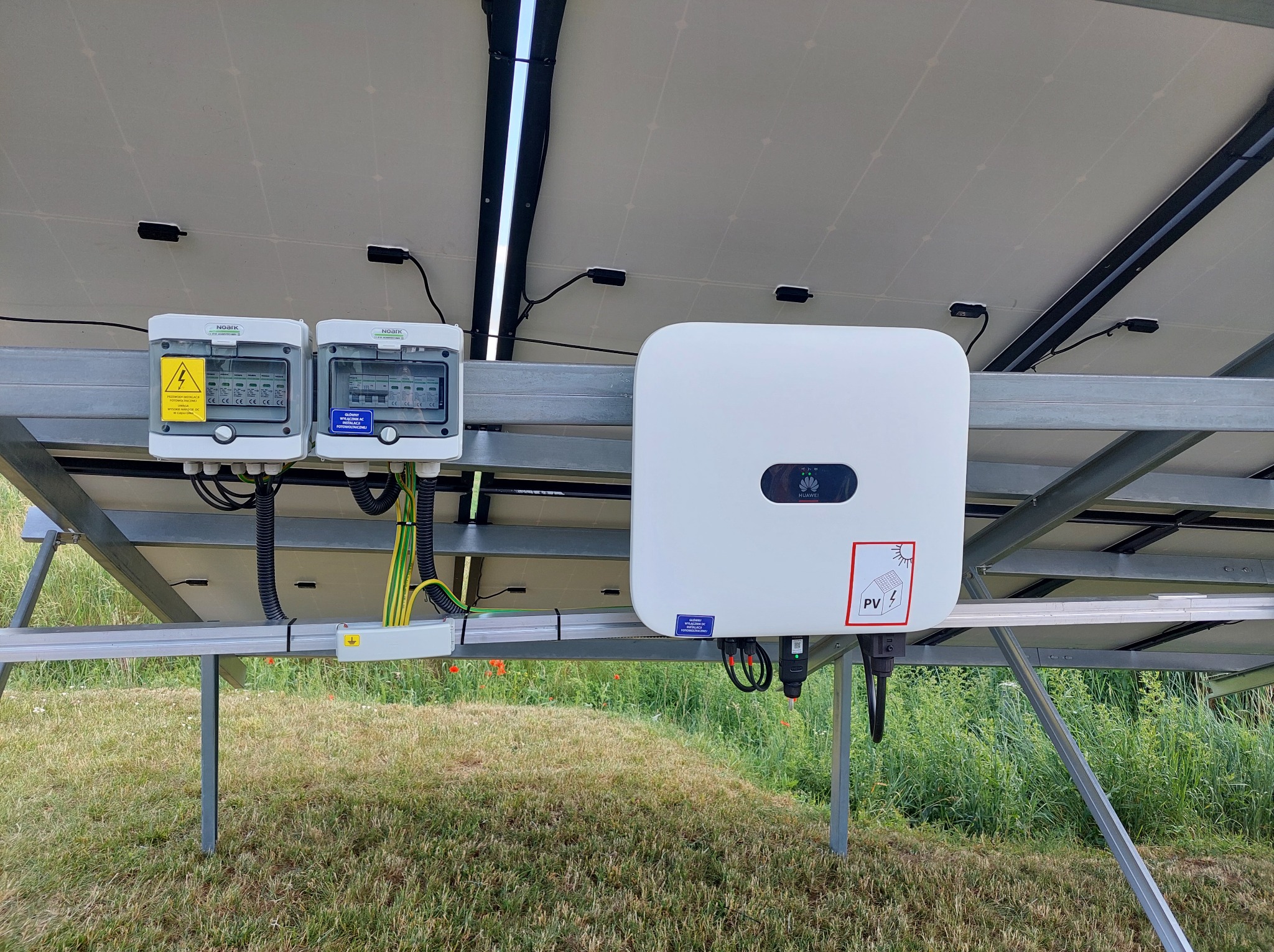 Instalacja fotowoltaiczna: widok od spodu na panele, biały inwerter Huawei i skrzynki z zabezpieczeniami. Przewody prowadzone w korytkach. Montaż na trawiastym terenie.