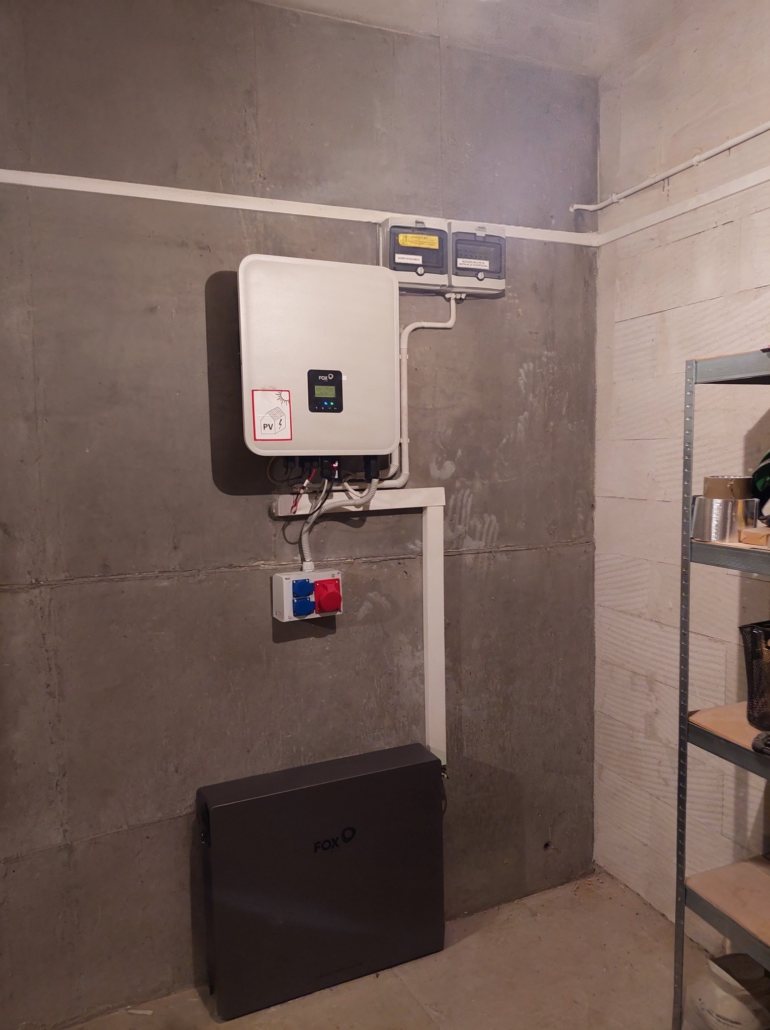 Kompletna instalacja fotowoltaiczna w Olsztynie. Widoczny inwerter, akumulator oraz zabezpieczenia na szarej, betonowej ścianie w pomieszczeniu.
