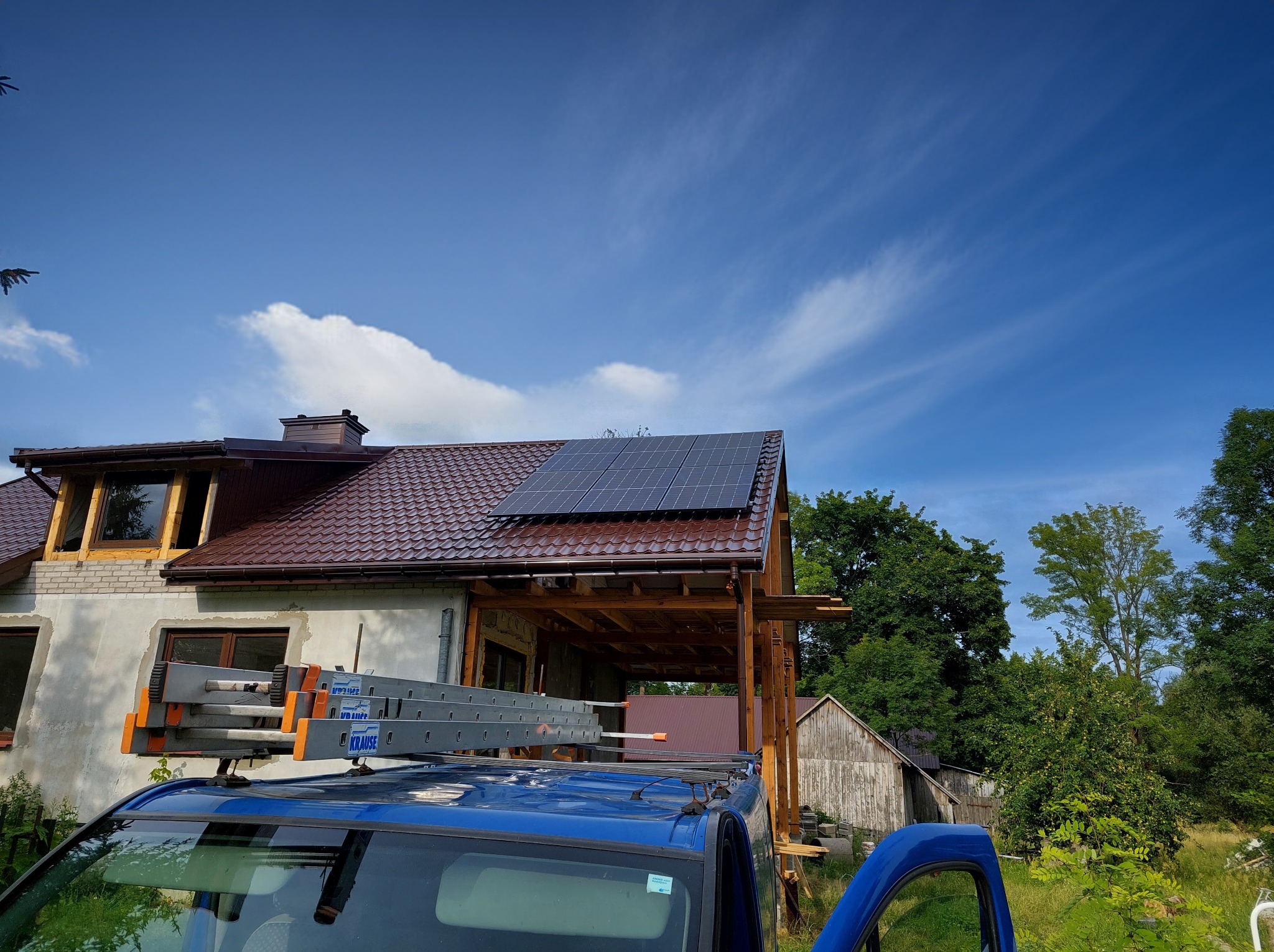 Dom z brązowym dachem i panelami fotowoltaicznymi w słoneczny dzień. Na dachu samochodu drabina Krause, sugerująca montaż lub serwis instalacji.