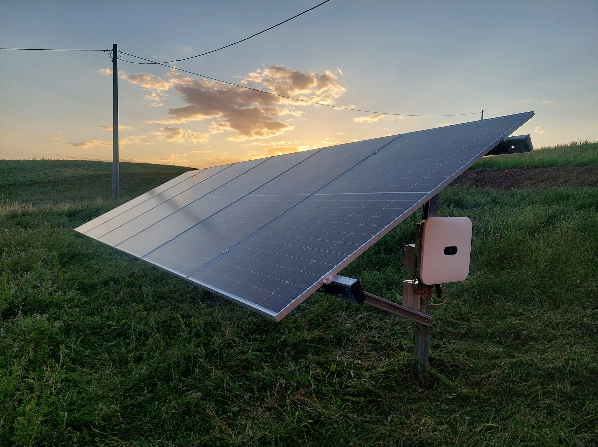 Panel fotowoltaiczny na łące, oświetlony zachodzącym słońcem. Widoczny biały inwerter i słup elektryczny w tle, delikatnie rozmyte chmury.
