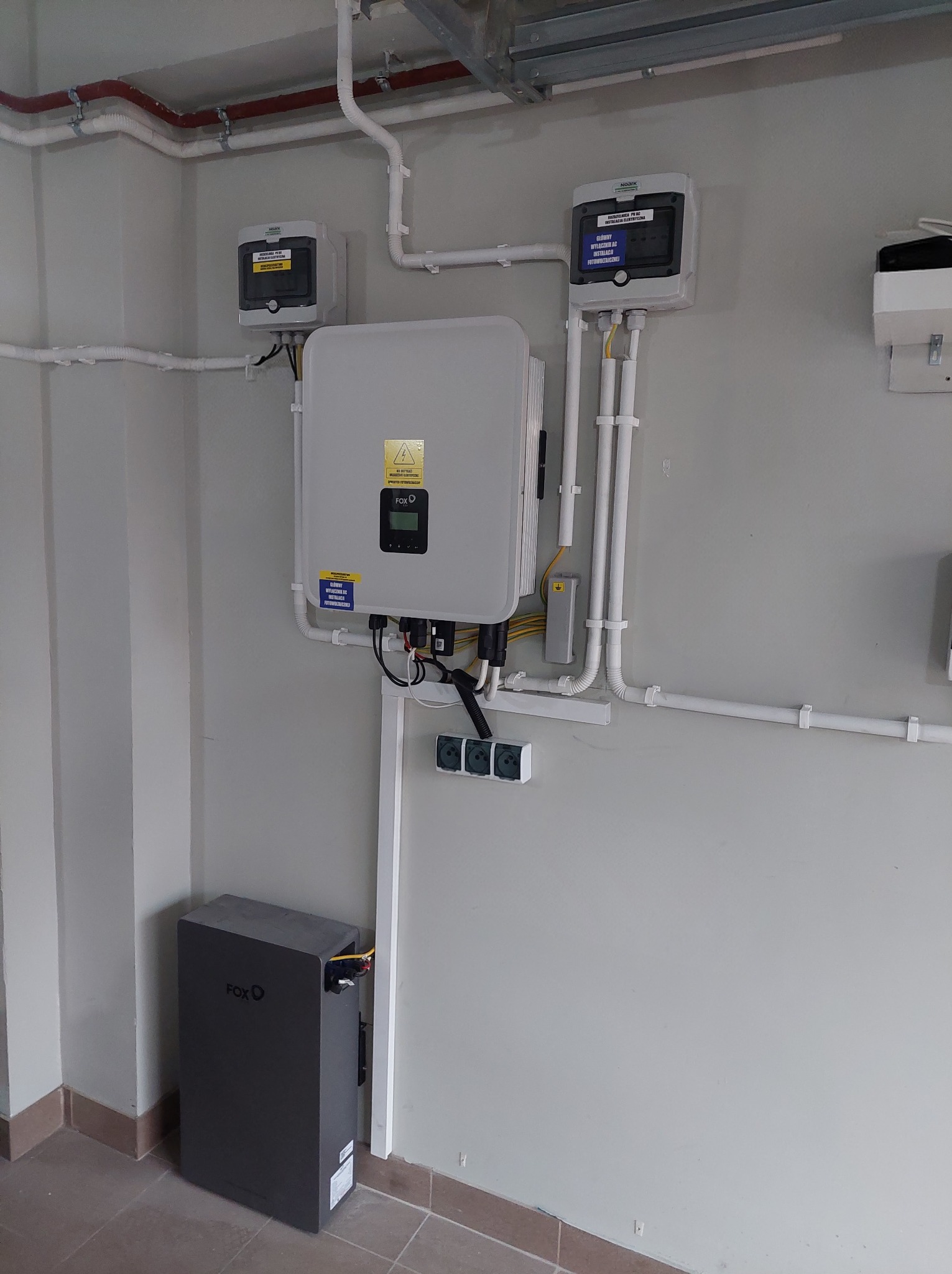 Zestaw fotowoltaiczny w pomieszczeniu: inwerter FoxESS, akumulator energii, zabezpieczenia AC/DC, okablowanie w białych peszlach. Widok na szarą ścianę i brązową podłogę.