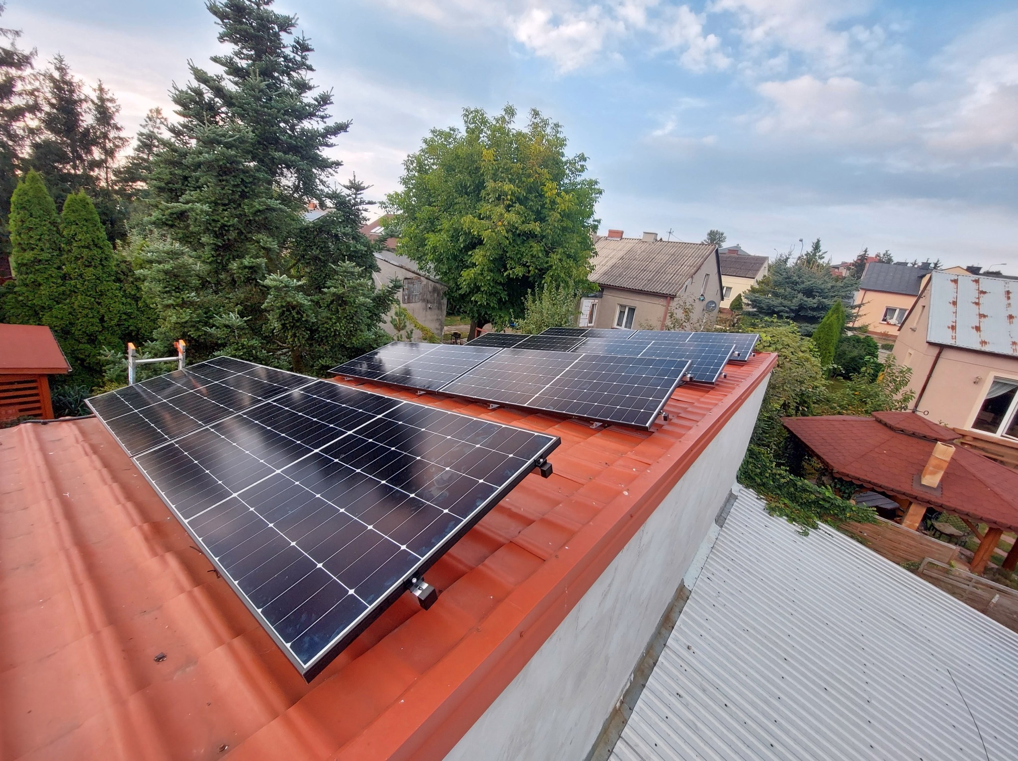 Panele fotowoltaiczne na czerwonym dachu budynku jednorodzinnego, widok z góry na tle zieleni i zabudowy mieszkalnej w słoneczny dzień.