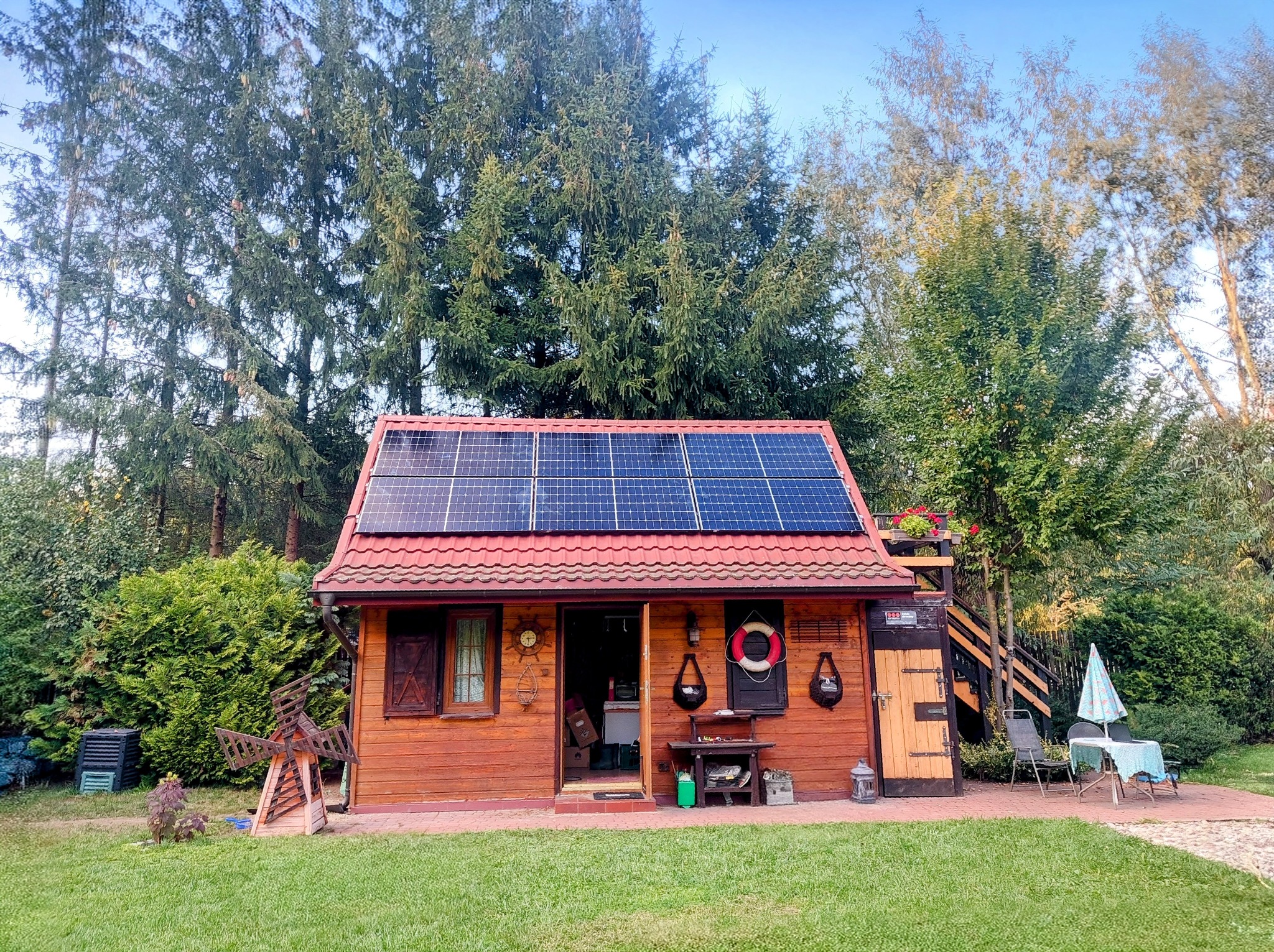 Drewniany domek z czerwonym dachem i panelami słonecznymi na tle zielonych drzew. Na ścianie koło ratunkowe i zegar. Widoczny stolik i krzesła na zewnątrz.