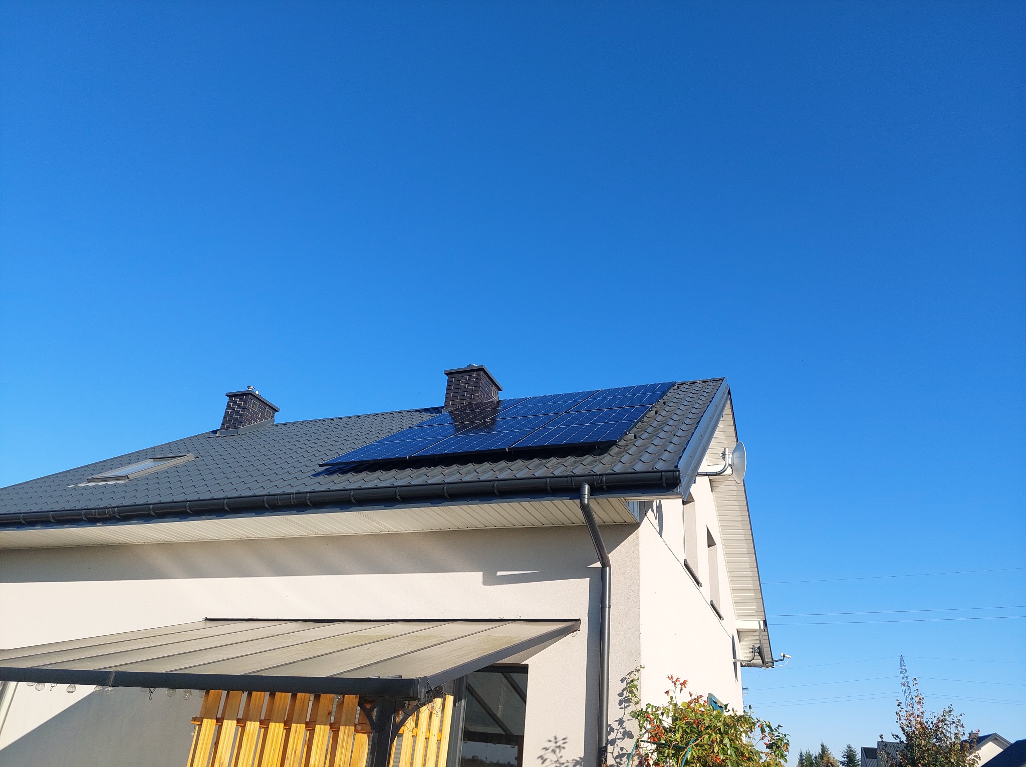 Panele słoneczne na dachu domu jednorodzinnego z szarą dachówką i dwoma kominami w słoneczny dzień. Widok z dołu na instalację fotowoltaiczną.