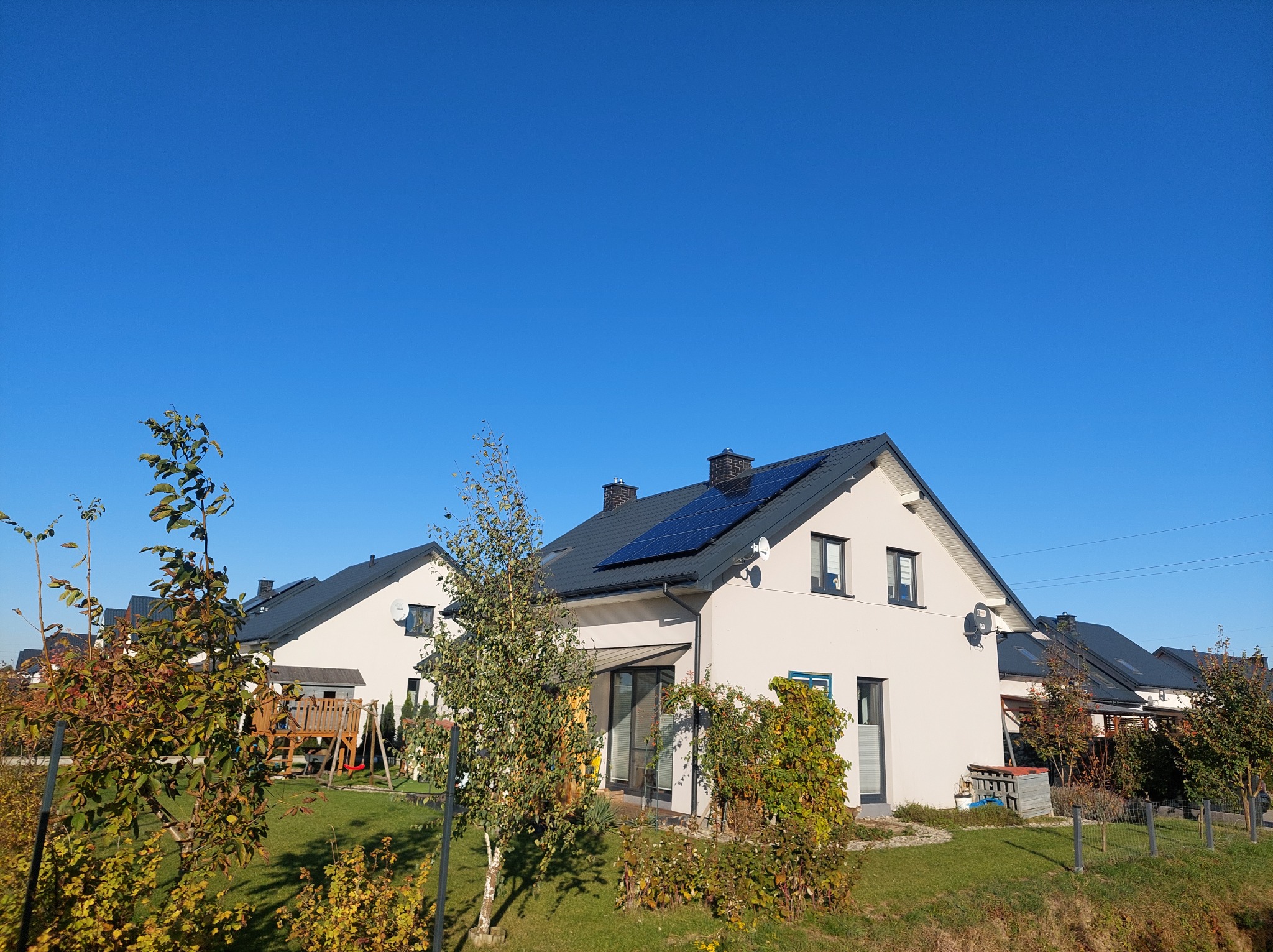 Biały dom jednorodzinny z panelami słonecznymi na dachu, widok z ogrodu w słoneczny dzień. Nowoczesna instalacja fotowoltaiczna na tle błękitnego nieba.