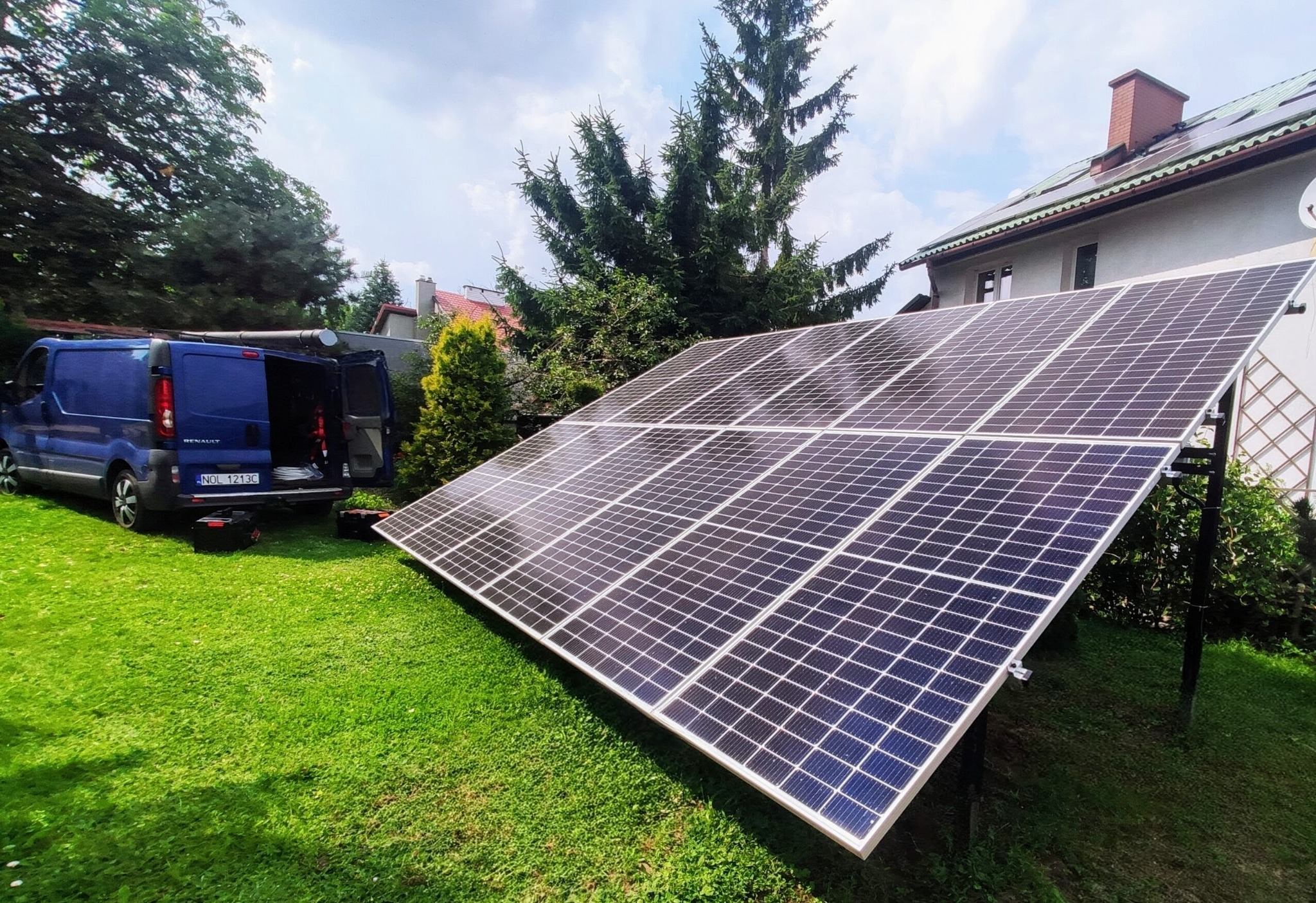 Instalacja fotowoltaiczna na trawie przed domem, z niebieskim vanem Renault z otwartymi drzwiami w tle, sugerująca mobilny serwis. Słoneczny dzień.
