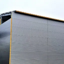 Montaż szarej płyty warstwowej na hali stalowej z użyciem podnośnika koszowego, widoczny pracownik w koszu i fragment logo firmy TOP HALE.