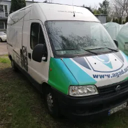 Biały bus marki Fiat z zielono-niebieskim logo firmy na tle trawy i budynków, z widoczną tablicą rejestracyjną PL 16429.