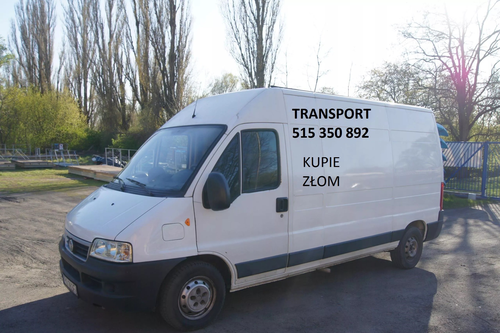 Biały furgon z napisem 'TRANSPORT 515 350 892 KUPIE ZŁOM' na tle drzew i metalowego ogrodzenia.