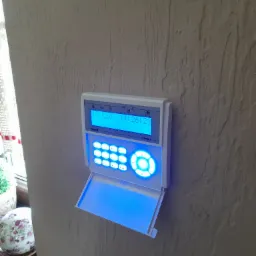 Panel alarmowy Satel Integro z niebieskim podświetleniem klawiatury i wyświetlacza, zamontowany na beżowej ścianie o wyraźnej fakturze, widoczny częściowo dekoracyjny element w tle.