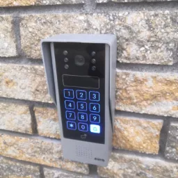 Szary wideofon z podświetlaną klawiaturą numeryczną, zamontowany na elewacji z cegły klinkierowej, widoczne logo producenta Eura.