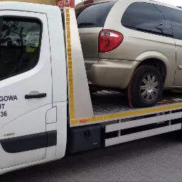 Laweta z załadowanym złotym minivanem na tle budynków mieszkalnych; na drzwiach lawety widoczny napis 'Pomoc Drogowa Skup Aut' i numer telefonu.