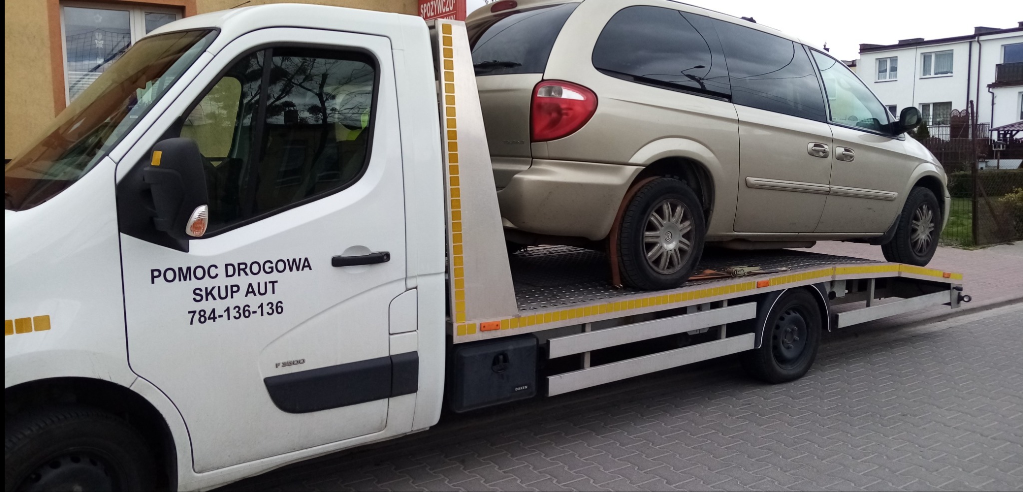 Laweta z załadowanym złotym minivanem na tle budynków mieszkalnych; na drzwiach lawety widoczny napis 'Pomoc Drogowa Skup Aut' i numer telefonu.