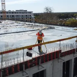Pracownik w pomarańczowej kurtce ustawia tachimetr na budowie, widoczny budynek w tle i dźwig.