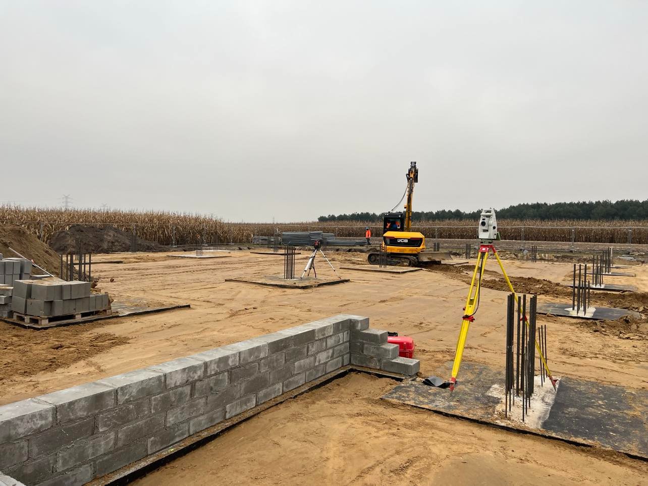 Prace ziemne na placu budowy z widocznym żółtym niwelatorem na statywie, fundamentami w ziemi, częściowo wymurowanym murem z bloczków betonowych oraz małą koparką w tle.