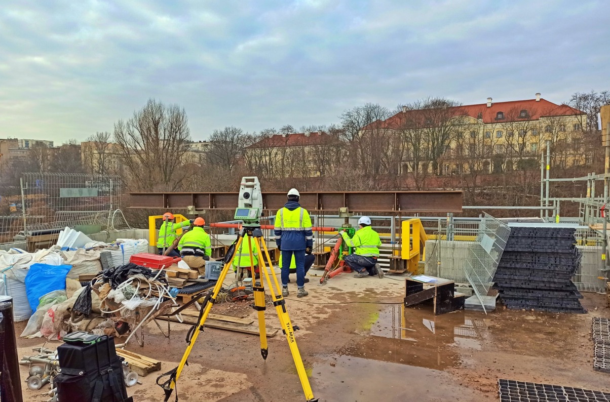 Praca na budowie mostu: zespół w odblaskowych kamizelkach używa teodolitu na trójnogu do pomiarów geodezyjnych; w tle budynek z czerwoną dachówką i stalowe elementy konstrukcyjne.