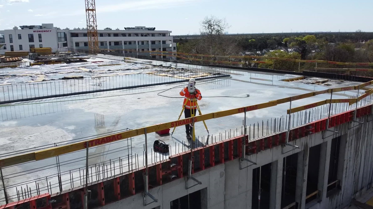 Pracownik w pomarańczowej kurtce ustawia tachimetr na budowie, widoczny budynek w tle i dźwig.