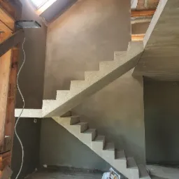 Wnętrze domu w trakcie wykańczania: surowe ściany pokryte świeżym tynkiem, drewniana konstrukcja dachu z oknem dachowym wpadającym światłem, betonowe schody bez wykończenia, widoczne instalacje...