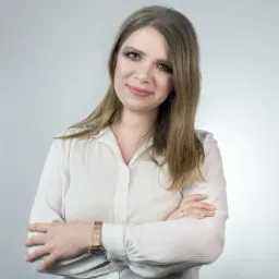 Portret kobiety z blond włosami, ubrana w białą koszulę, z zegarkiem na ręce, skrzyżowanymi ramionami i delikatnym uśmiechem na jasnym tle.