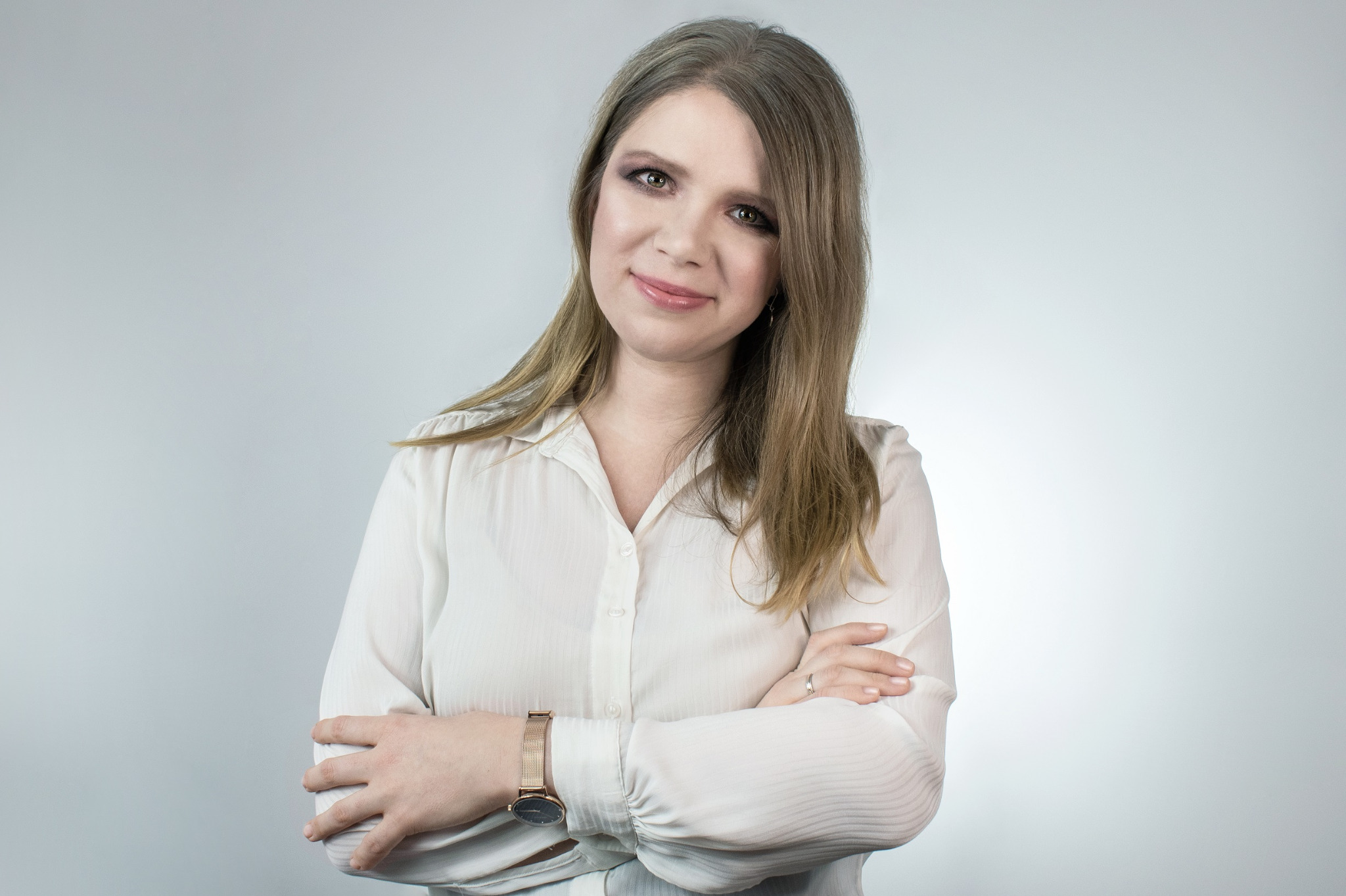 Portret kobiety z blond włosami, ubrana w białą koszulę, z zegarkiem na ręce, skrzyżowanymi ramionami i delikatnym uśmiechem na jasnym tle.