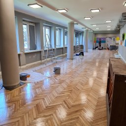 INSAJ-BUD Natallia Bahdanovich - Sala z odnowionym parkietem ułożonym w jodełkę, widoczne ślady prac remontowych, drabina i wiadro, kolumny wzdłuż ścian, okna zabezpieczone folią malarską.