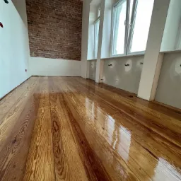 Lakierowana, odnowiona podłoga z desek w jasnym pomieszczeniu z ceglaną ścianą i dużymi oknami.