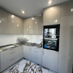 Nowoczesna kuchnia z szarymi szafkami, marmurową podłogą i wbudowanym piekarnikiem oraz mikrofalówką, widoczne folie ochronne na frontach szafek.