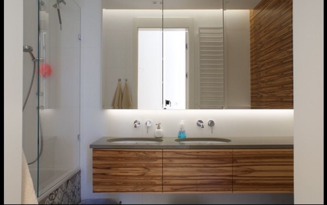 Nowoczesna łazienka z drewnianą szafką pod umywalką z dwoma okrągłymi umywalkami, oświetleniem LED pod lustrem i szklaną kabiną prysznicową z widoczną różową gąbką.