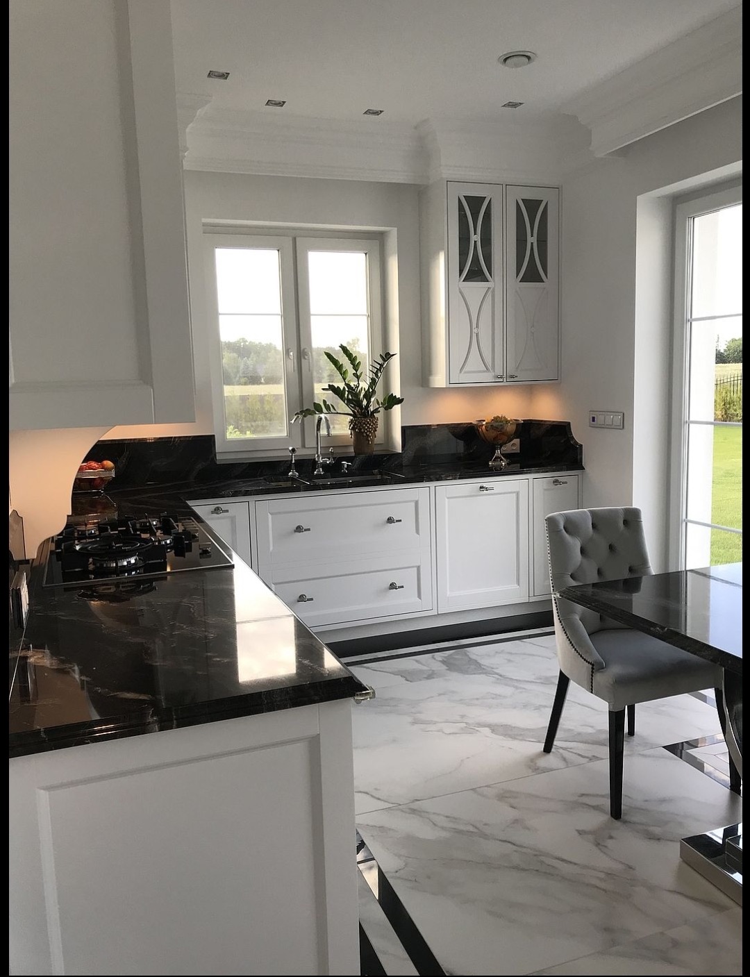 Elegancka, jasna kuchnia z czarnym, błyszczącym blatem, białymi szafkami, marmurową podłogą i widokiem na zieleń za oknem.
