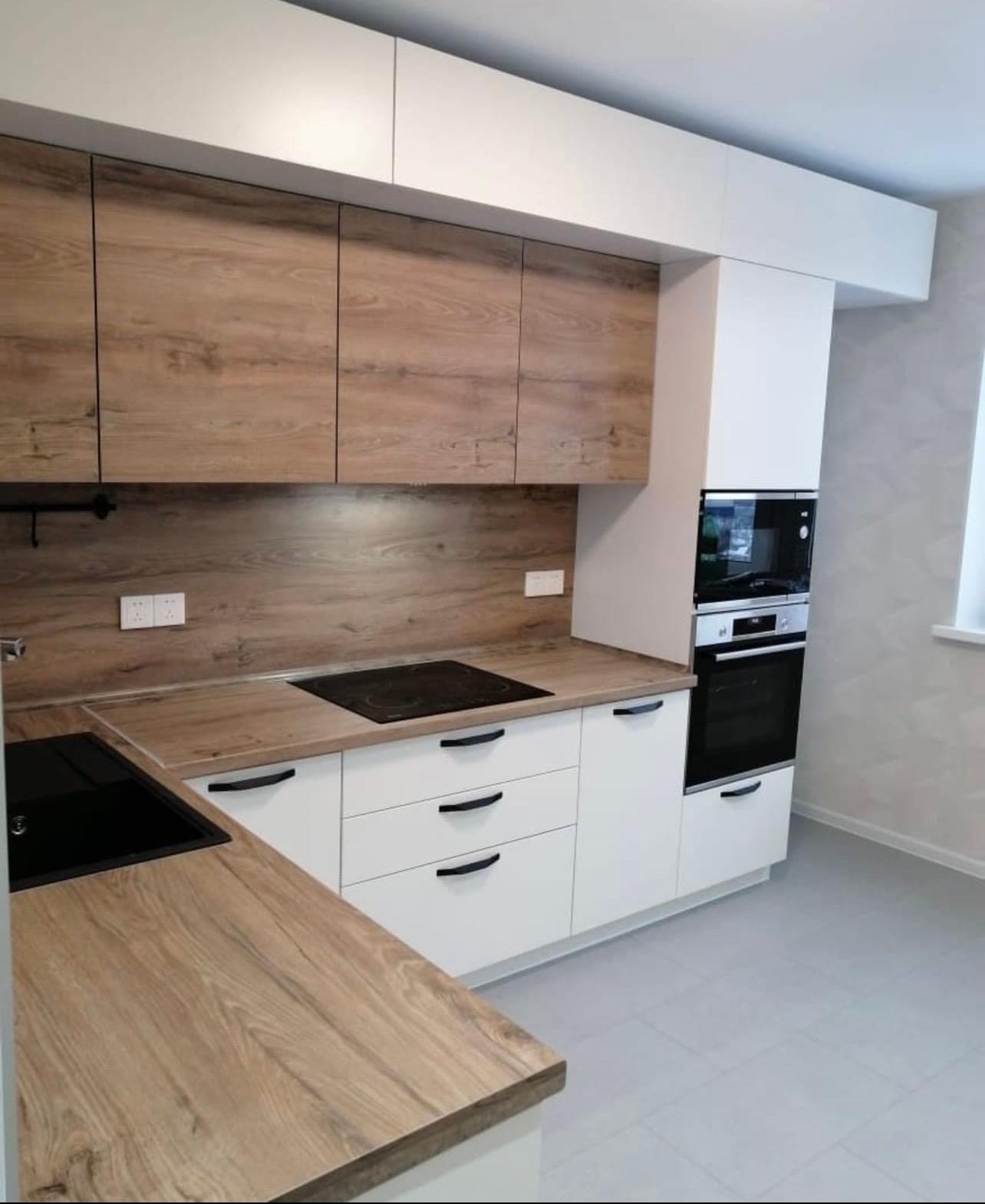 Nowoczesna kuchnia z białymi szafkami dolnymi i górnymi szafkami w kolorze drewna, czarny blat kuchenny i płyta indukcyjna, czarne uchwyty, piekarnik i mikrofalówka w zabudowie.