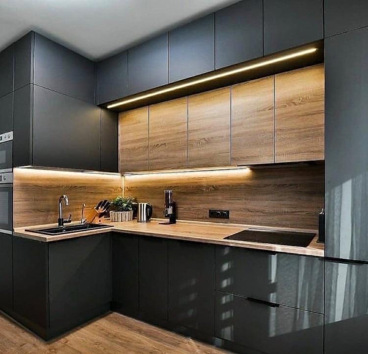 Nowoczesna kuchnia z ciemnymi, matowymi szafkami dolnymi i górnymi wykończonymi drewnem, oświetlona listwami LED pod szafkami, z czarnym zlewem narożnym i drewnianym blatem.