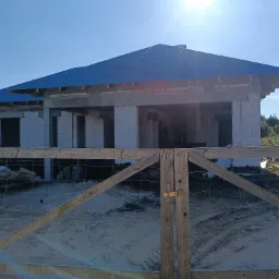 Niedokończony budynek jednorodzinny z niebieskim dachem, widoczny przez drewniane ogrodzenie z siatką, w słoneczny dzień.