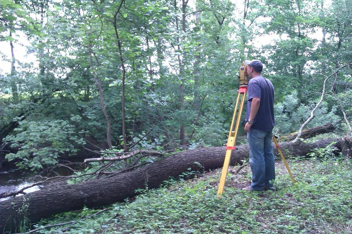 Geodeta w terenie leśnym, używający niwelatora na żółtym statywie, obok powalonego pnia drzewa i małego strumienia.