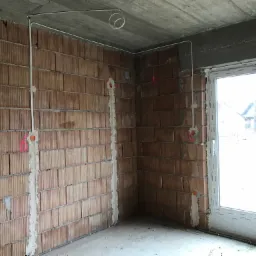 Surowy mur z cegły z wyfrezowanymi bruzdami wypełnionymi gipsem maskującymi przewody elektryczne, widoczne puszki instalacyjne, kabel zwinięty w pętlę pod sufitem i okno PCV w tle.