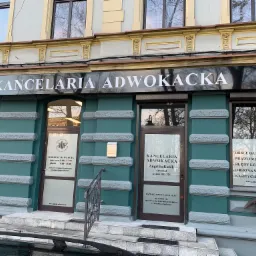 Fasada budynku kancelarii adwokackiej z szyldem 'Kancelaria Adwokacka', oknami z informacjami o usługach i godzinach otwarcia, oraz wejściem z kilkoma schodami.