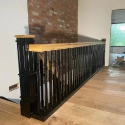 Nowoczesna balustrada z czarnymi, pionowymi prętami i jasnym, drewnianym poręczą, na tle ściany z cegły i drewnianej podłogi, widok z dołu.