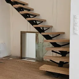 Ażurowe schody z drewnianymi stopniami i czarną metalową konstrukcją wspornikową, widoczne podczas montażu, z niezabezpieczonymi przewodami na ścianie i lustrem w tle.