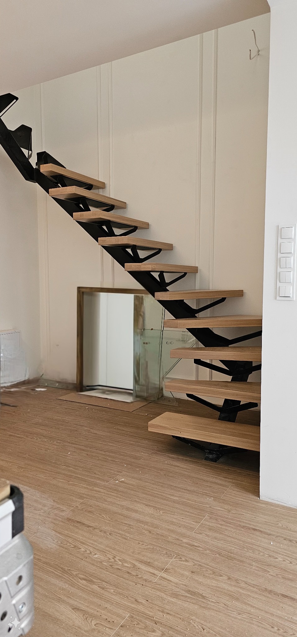 Ażurowe schody z drewnianymi stopniami i czarną metalową konstrukcją wspornikową, widoczne podczas montażu, z niezabezpieczonymi przewodami na ścianie i lustrem w tle.