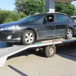 Czarny Peugeot kombi na platformie lawety z otwartymi drzwiami, na stacji benzynowej.