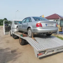Srebrny samochód osobowy marki Audi A4 na platformie lawety, z otwartymi drzwiami kabiny i widocznymi tablicami rejestracyjnymi, w tle plac budowy z koparką.